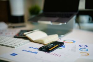 Mesa de trabalho com notebook, calculadora, teclado, caderno aberto e gráficos financeiros impressos sobre a mesa.
